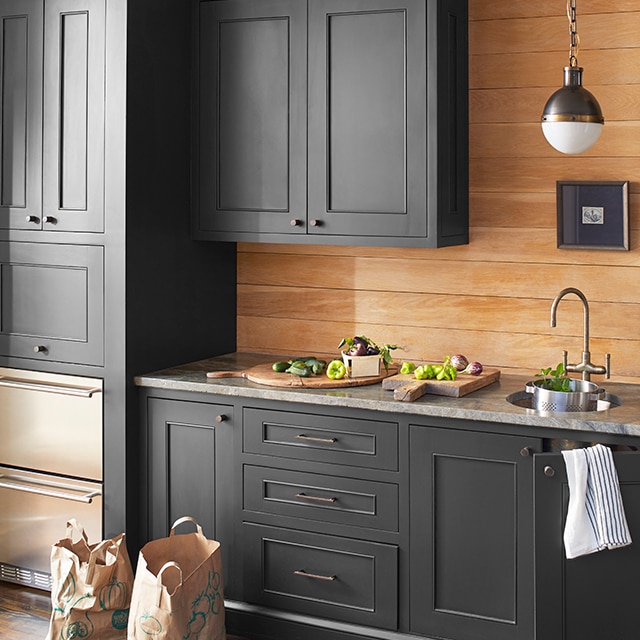 A section of a pretty kitchen with dark gray painted cabinets, granite countertops strewn with fresh produce, wood shiplap walls, and paper grocery bags on the wood floor.