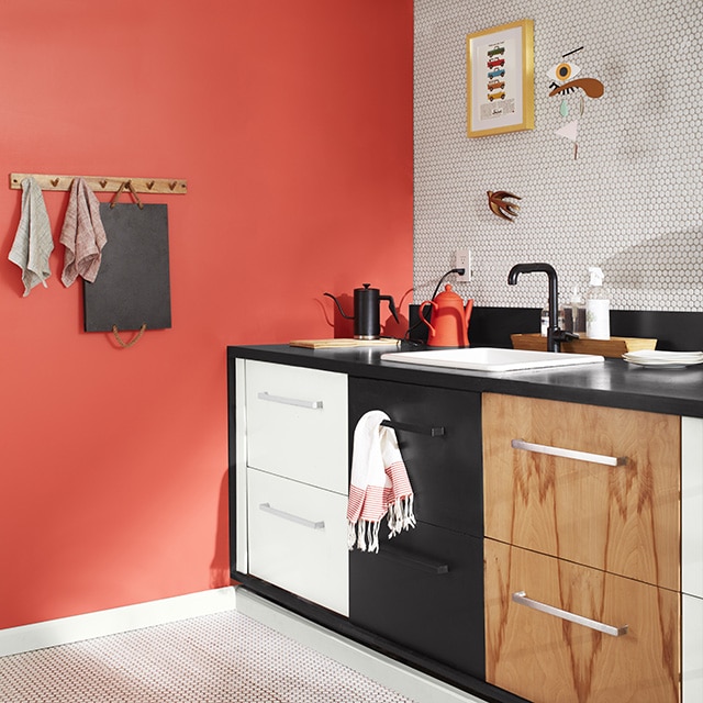 A sleek kitchen corner with a coral painted wall, a white tiled backsplash wall and floor, white upper wall and exposed pipes, and three sets of drawers: one painted white, the second black, and the third woodgrain.