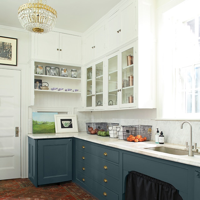 A corner of a white-painted kitchen with open shelving, a crystal and gold chandelier, printed red rug, and dark teal blue lower kitchen cabinets.