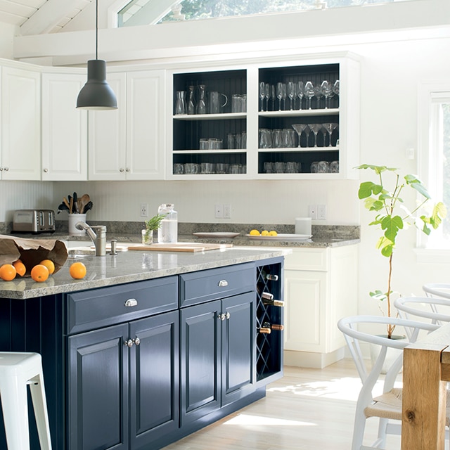 A bright, open white-painted kitchen with white cabinets, vaulted ceiling and beams, a beautiful dark blue painted island and gray countertops.