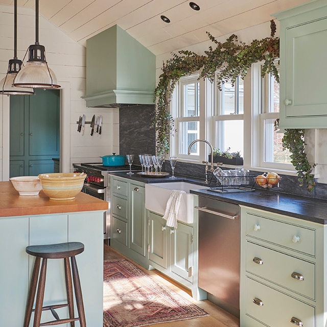 An airy, welcoming kitchen with light blue-green painted cabinets and island, white walls and a white shiplap ceiling, and a red area rug on the sunlit wood floor.