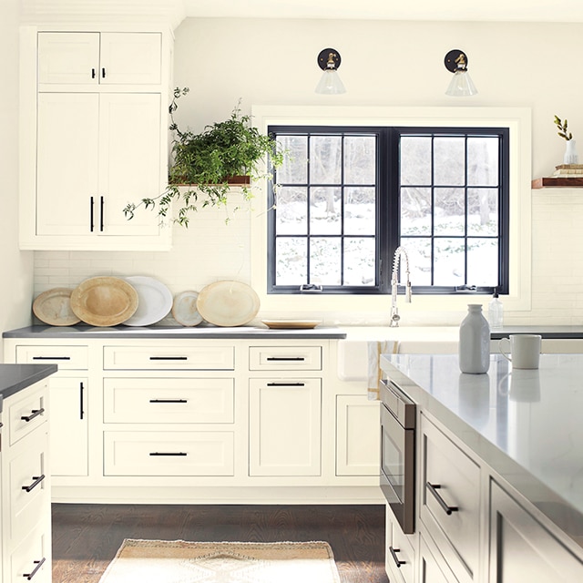 A large sunny kitchen with off-white-painted walls and cabinets, a print area rug, and a window above the sink with black-painted window trim.