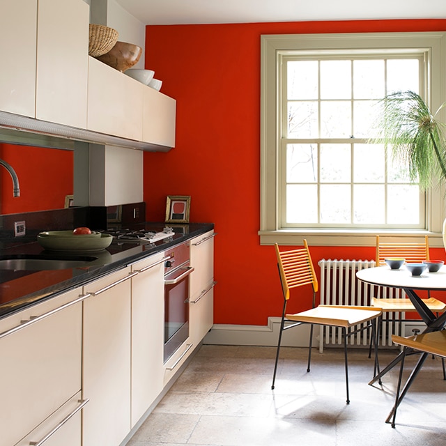 Cuisine étroite avec petite table ronde et chaises, armoires et murs blanc cassé, grande fenêtre et mur d’accent d’un flamboyant rouge orangé.