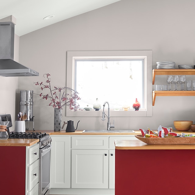 A cheery small kitchen with lavender-gray walls, red-painted cabinet siding, white-painted lower cabinets, wooden floors and a sunny bright window above the sink.