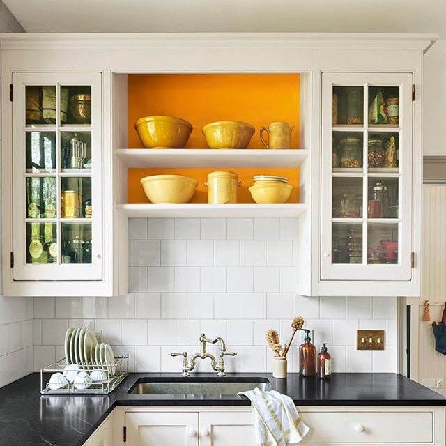 A mostly white-painted kitchen with black countertops and orange accent behind shelving.