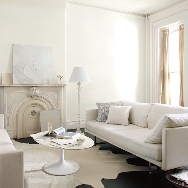 Neutral living room with white walls and an off white wall mantel, and a white circle coffee table in between two cream couches.