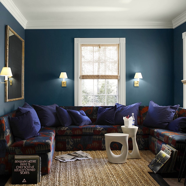 A comfy living room with dark, rich blue painted walls, white ceiling and trim, a multi-colored wrap-around sofa with blue pillows, modern white tables, and a seagrass rug.