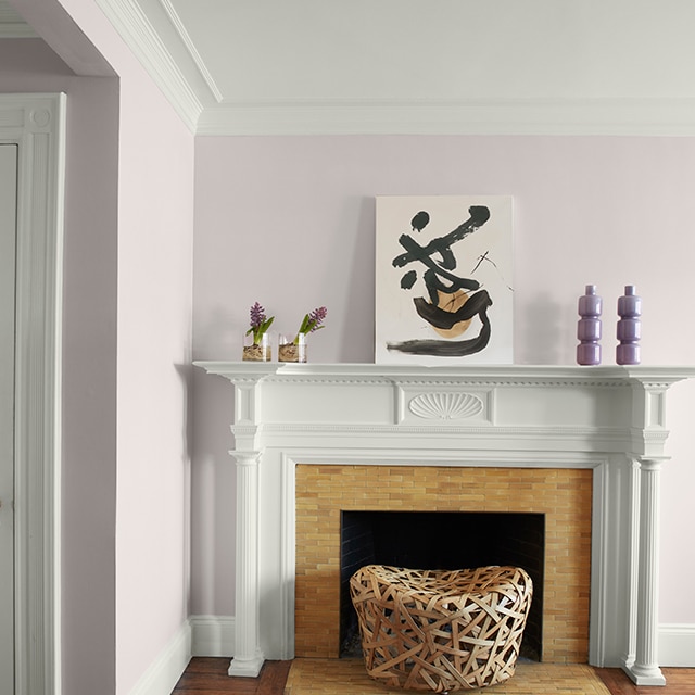 Pretty light violet-painted corner walls, white crown molding, trim and fireplace mantel with modern artwork and purple vases, and a white hallway door on the left. 