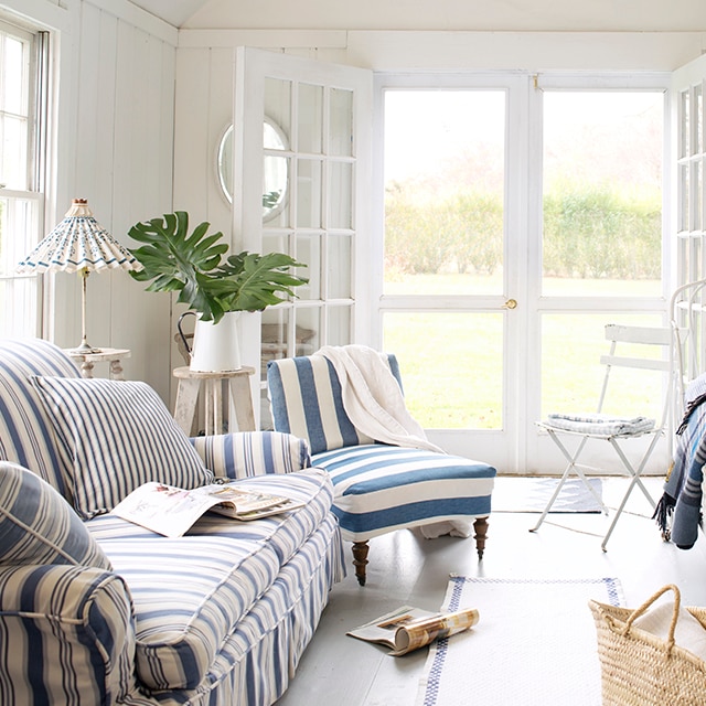 This bright, white-painted cottage living room has a relaxed, coastal vibe with blue and white stripe and slipcovered furniture, a vaulted ceiling, open French doors and screen doors.