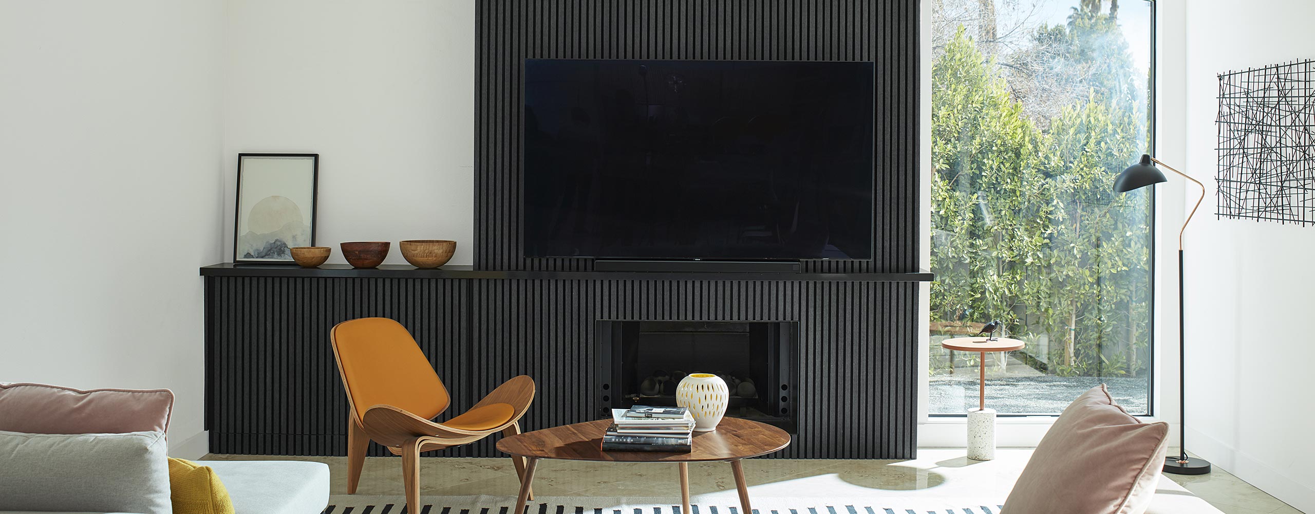 A mid-century modern style living room with bright, white-painted walls and vaulted ceiling, and a black paneled accent wall, built-in cabinet and fireplace, and large screen tv.