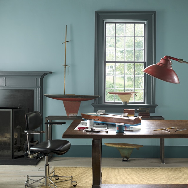 A spacious home office with blue-gray painted walls, a dark gray mantel and trim, a large wood desk, leather swivel chair, red floor lamp, and wooden toy boats.