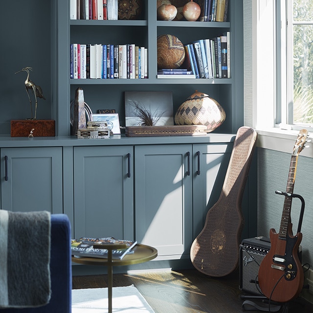 Sunlight reflecting on beautiful gray-blue painted built-in shelves and cabinets in a home office, with a guitar, amp, and case against the lower right wall.