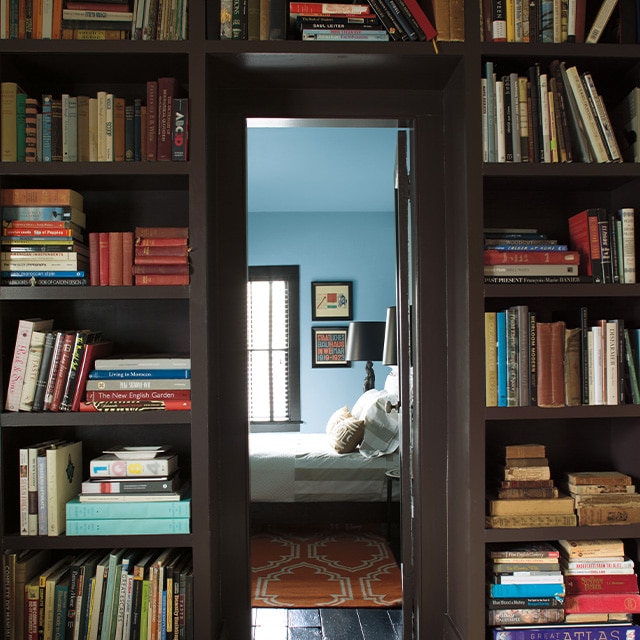 A doorway framed by dark brown painted built-in bookshelves open into a blue painted bedroom.