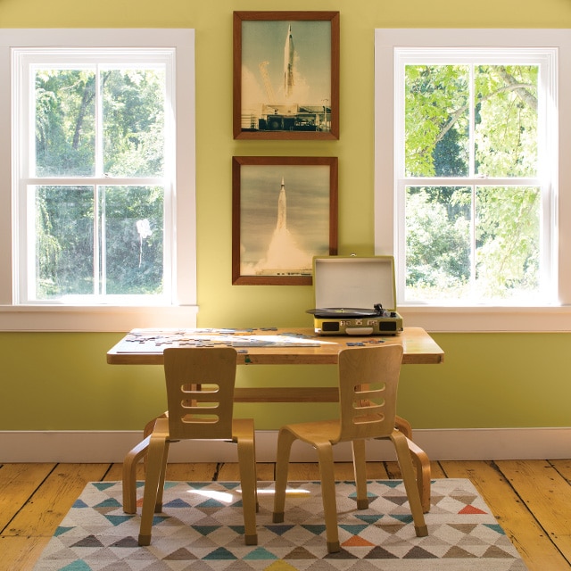 A children’s wooden desk, two chairs, and a multicolor rug in front of a light green painted wall with white trim and rocket pictures flanked by two windows.