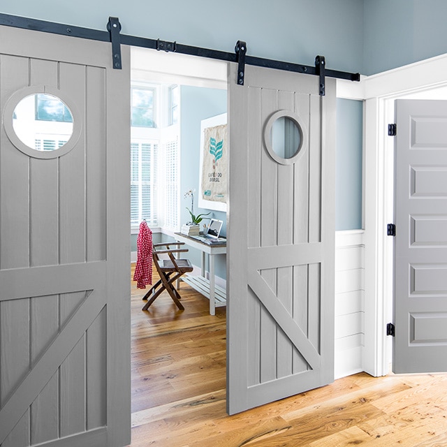 A pretty, blue painted hallway looking into a blue painted office space with a small white desk, a brown chair, white trim, and open gray painted sliding barn doors.