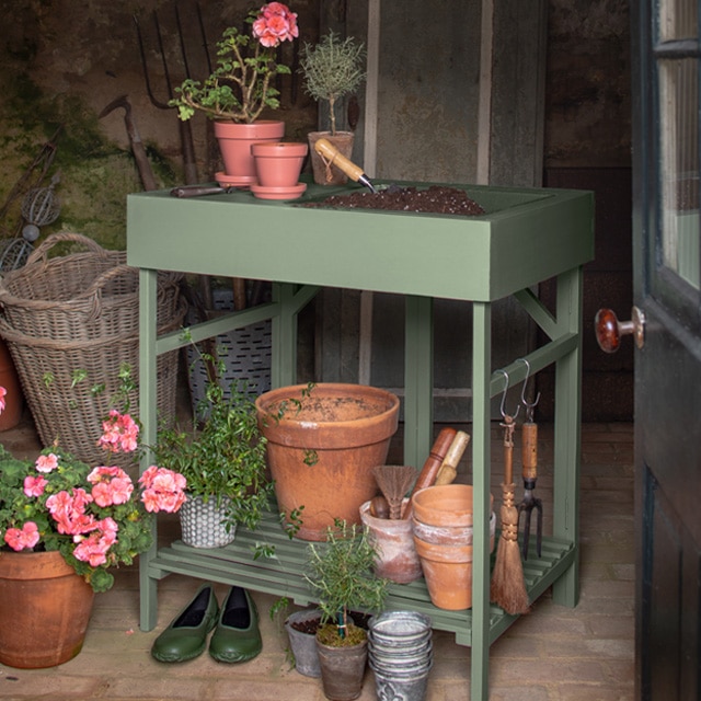 A mossy green-painted gardening stand with clay pots, pink flowers, gardening tools, and baskets adds to a cheery workspace inside a shed. 