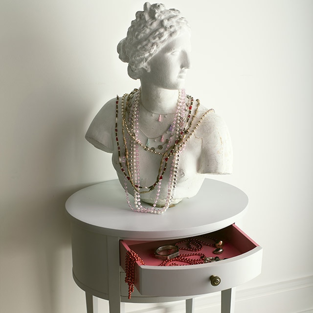 A carved bust with jewellery draped over it on a light-gray-painted round table with an open scarlet red-painted interior drawer in front of a white-painted wall.