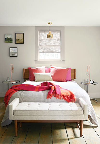 A relaxing bedroom with light gray walls, full size bed with pink pillows and a throw blanket, and cream bench.