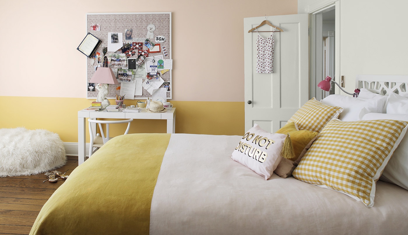 Une chambre à coucher peinte en rose et jaune arborant un fauteuil poire, un tableau de liège, un bureau blanc et un couvre-lit à deux tons.