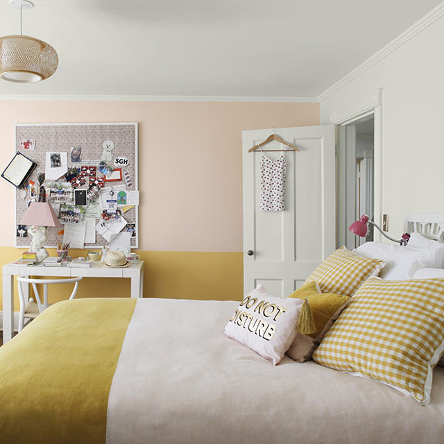 Une chambre à coucher peinte en rose et jaune arborant un fauteuil poire, un tableau de liège, un bureau blanc et un couvre-lit à deux tons.