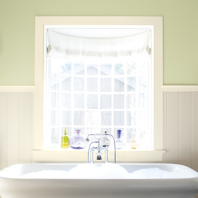 Une salle de bains avec des lambris peinturés dans une nuance de blanc, la partie haute des murs enduite d’une couleur verte et une baignoire blanche sous une baie vitrée.