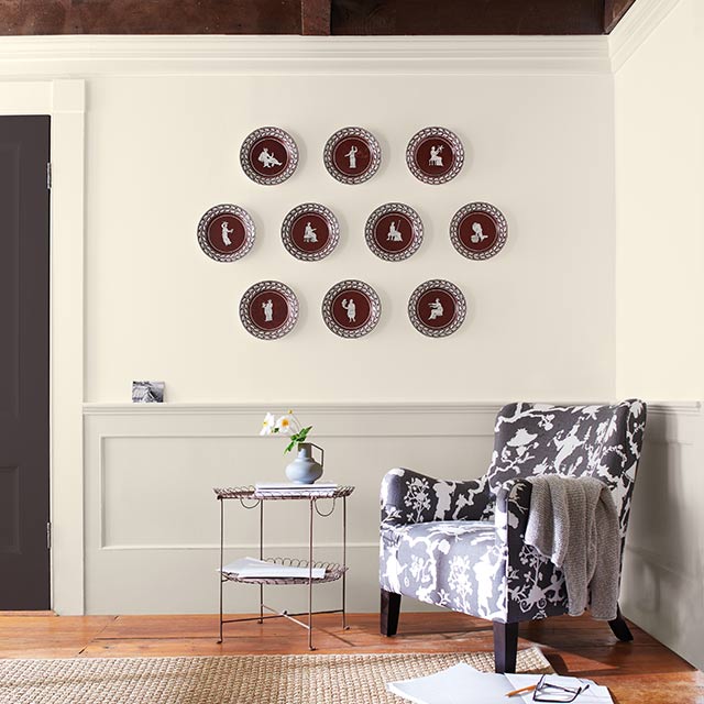 Un coin salon avec des murs peints en blanc et des lambris gris, des moulures et un plafond avec des poutres en bois, une porte brune, une chaise mouchetée et des plats décoratifs.