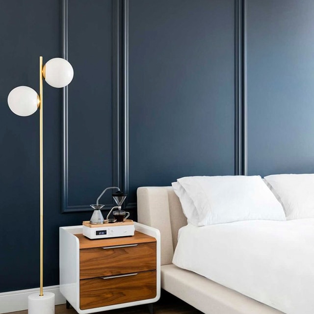 A contemporary bedroom with dark blue-painted walls, white trim, a cream-coloured platform bed, a modern nightstand, and a standing lamp with two round, oversized lightbulbs.