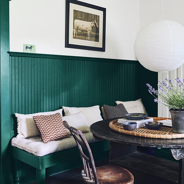A charming kitchen table nook with green-painted wainscotting and bench, white upper wall, wooden table and chair, and potted lavender beneath a contemporary globe chandelier.