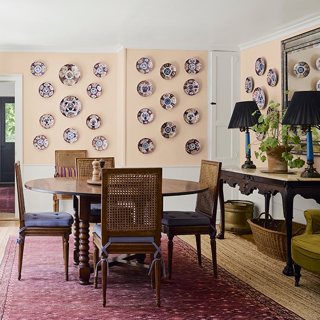Salle à manger rose pêche décorée d'assiettes décoratives sur les murs avec table en bois, quatre chaises, table d'appui et tapis bourgogne sur plancher de bois franc clair.