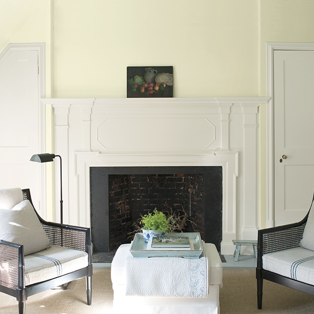 A pretty yellow-green painted wall with a white fireplace flanked by two white doors, with two chairs and a white ottoman in front.