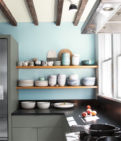 A residential kitchen with two exposed wooden shelves showcase bowls and plates against a light blue colored wall.
