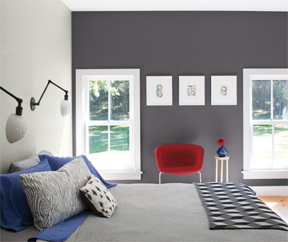 A bedroom in varied gray tones features a contemporary red accent chair and two windows with a view of outdoor greenery.