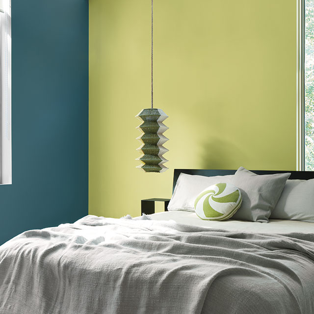A bedroom with two painted walls, one in a soft yellow-green, and the other in an aquatic deep blue, and a bed with gray bedding, and a green and white accent pillow.