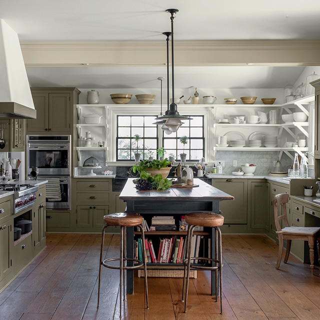 An inviting, open kitchen with sage green-painted cabinets, white open shelving with a collection of white stoneware, a rustic wooden center island and wood floor.