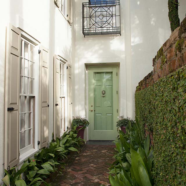 The front entrance of an elegant, white-painted house with beige shutters, a light-green front door, brick walkway and brick wall covered in foliage.