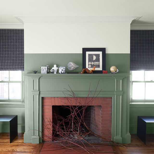 Pièce avec mur mi-blanc mi-vert, plafond blanc, manteau de cheminée vert, plancher de bois et deux fenêtres.