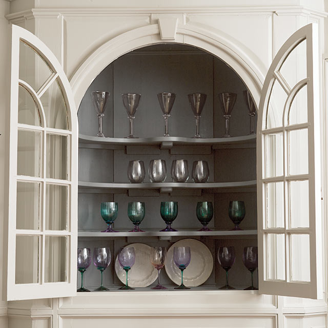 A light-gray-painted built-in hutch with open glass doors, holding coloured glassware and plates.