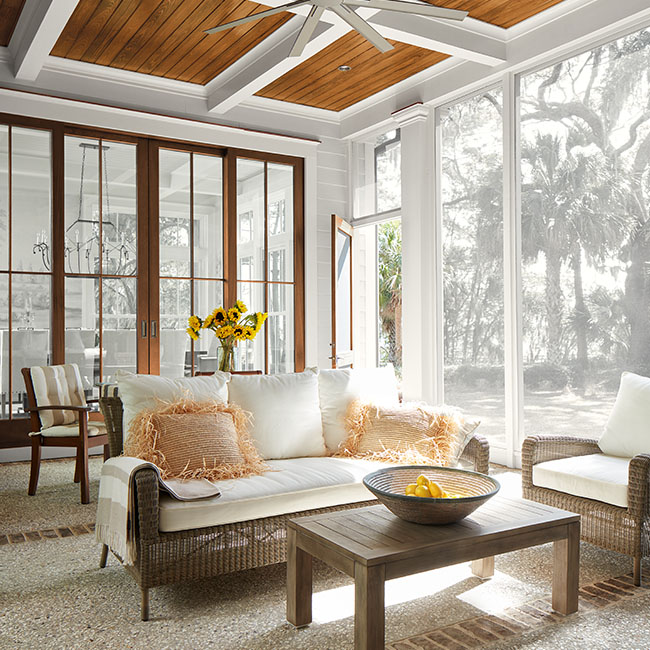 A beautiful screened-in porch with a wood paneled and white beamed ceiling, white trim, a ceiling fan and white cushioned furniture.
