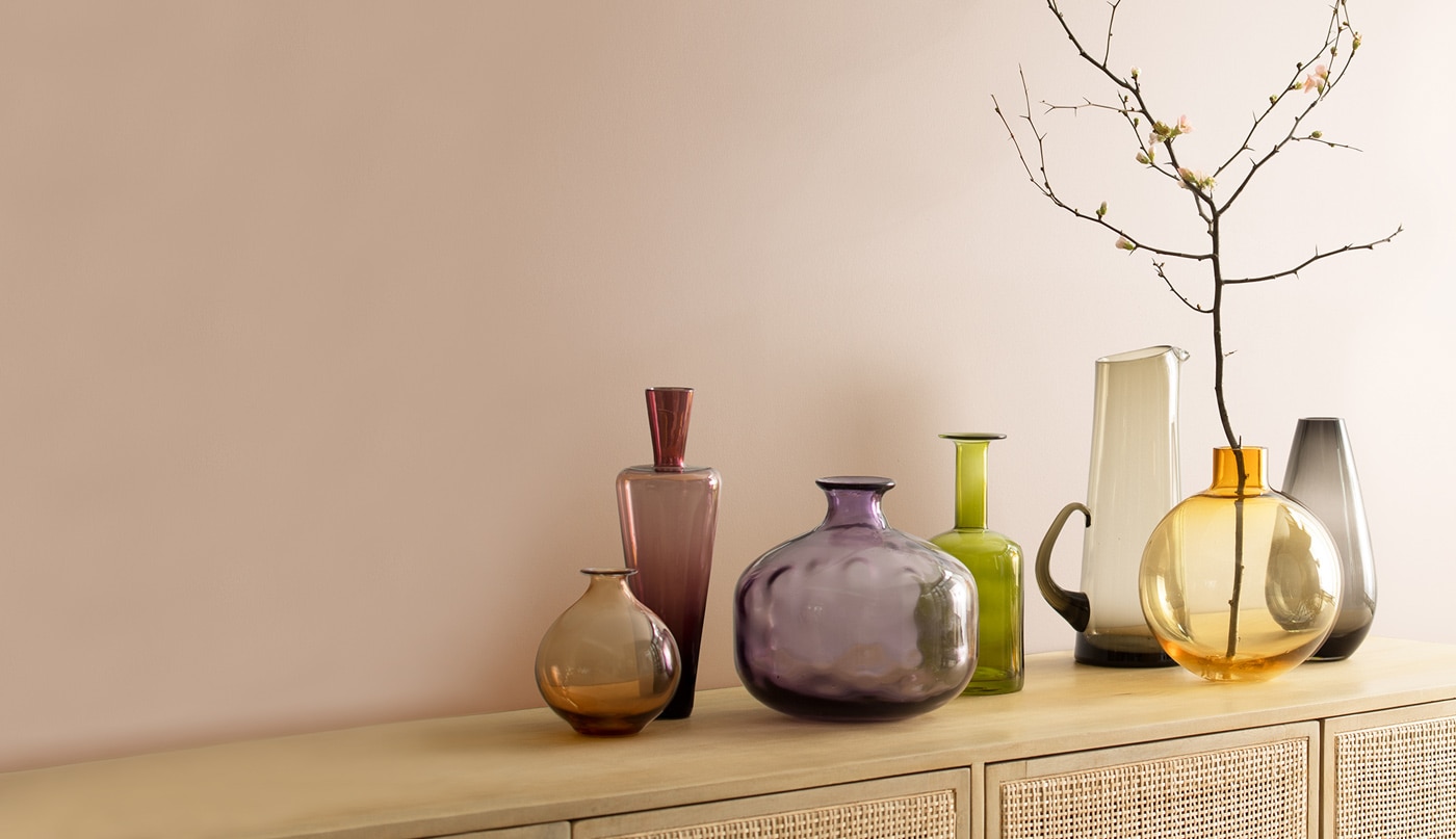 Dusty pink walls behind a rattan cabinet with an array of glass vases placed on top. 