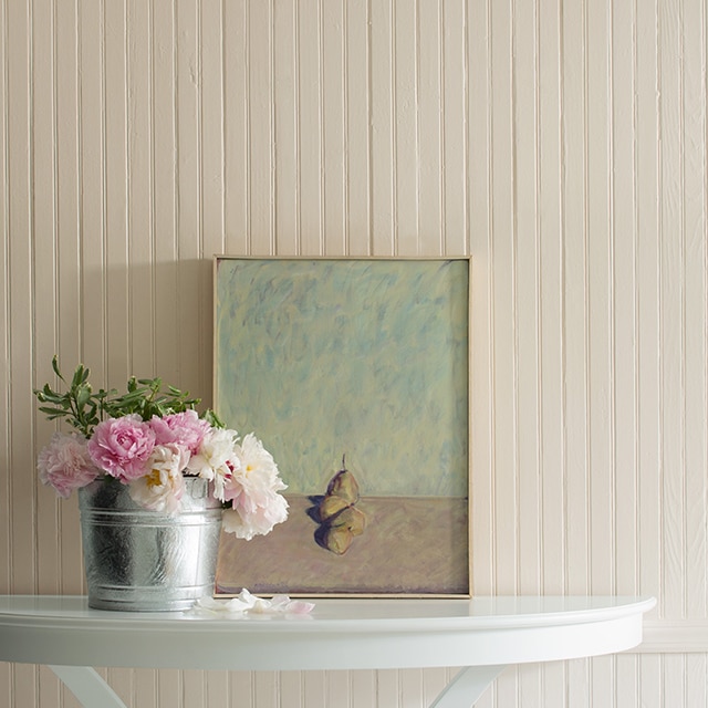 Pale pink walls with a painting and a tin bucket of pink flowers on top of a console.