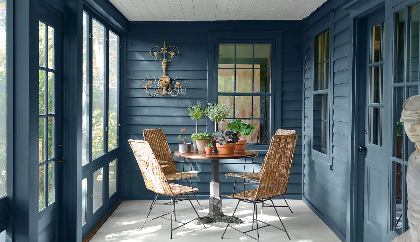 Enclosed blue-painted cabin porch with modern table, chairs, house plants, and wall light.