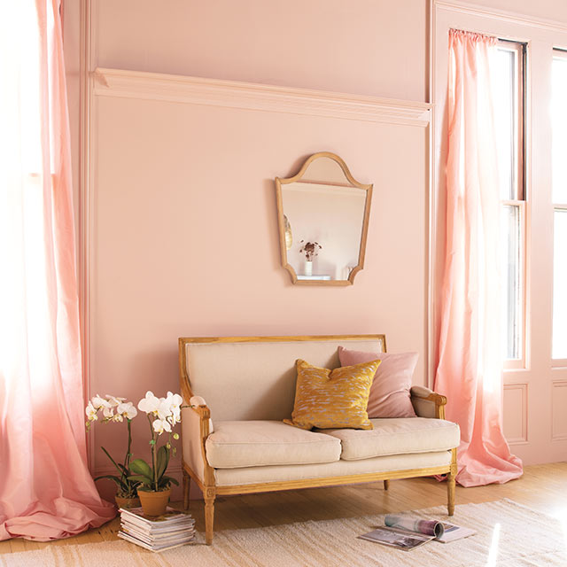 Un coin salon aux murs peints en rose corail arborant des moulures assorties et une causeuse blanc cassé au-dessus de laquelle est accroché un miroir.