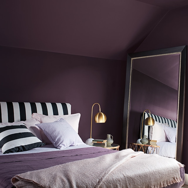 Chambre à coucher violet foncé avec moulures blanches, tête de lit et coussin à rayures noir et blanc, et couvertures mauves et blanches.