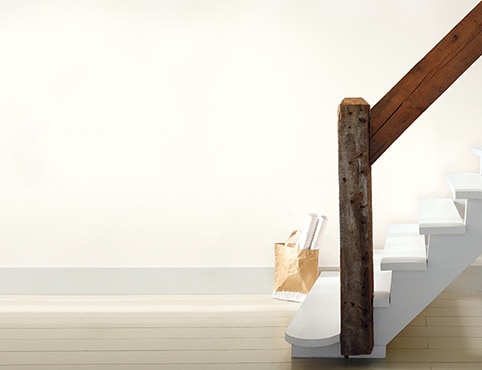 A rustic white staircase with wooden handrail in an off-white room; a tote filled with rolled posters sits on the floor. 