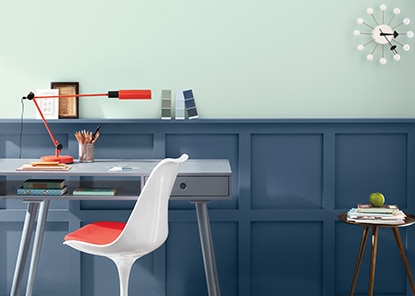 A room with dark blue painted wainscoting, a light blue wall, gray desk and white and red chair.