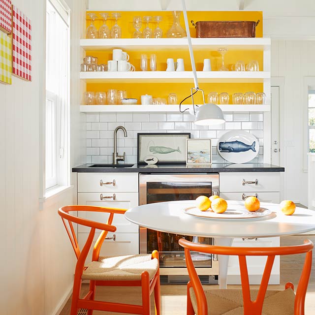 A white-painted kitchen with open shelving, a yellow accent wall, a white table and orange chairs.
