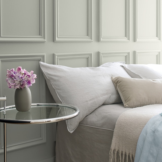 Moulure de couronne grise derrière un lit avec une literie grise et une table d’appoint en verre avec un petit vase de fleurs violettes.