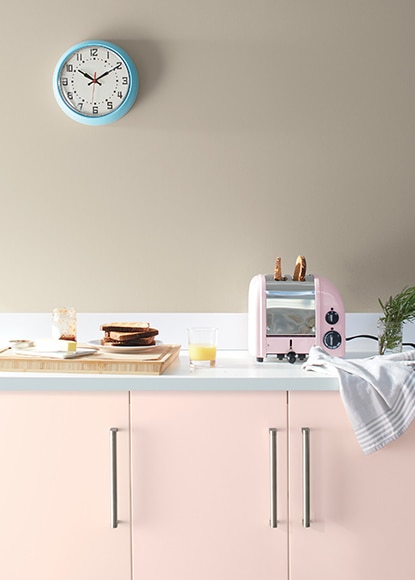 Les murs sont gris avec des armoires rose clair et des comptoirs blancs avec des appareils de cuisine et une petite horloge murale bleue.
