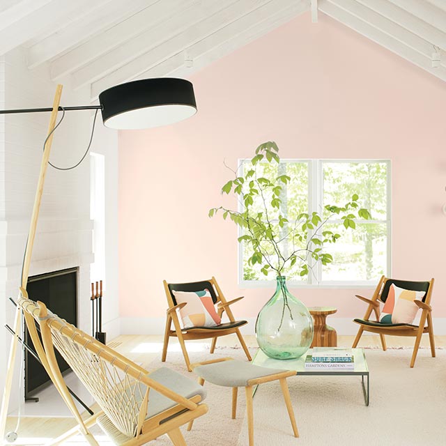 Un salon lumineux présentant un plafond à poutres apparentes blanches, un mur d’accent peint en rose clair, trois fauteuils et un canapé.