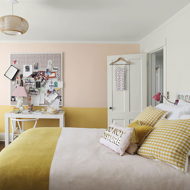 Une chambre à coucher peinte en blanc doux et jaune arborant un fauteuil poire, un tableau de liège en désordre, un bureau blanc et un couvre-lit à deux tons.